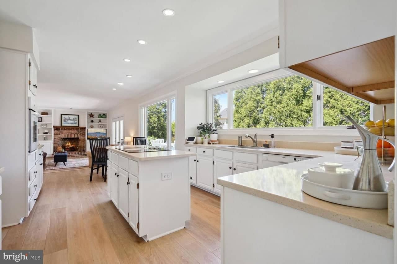 Bright white kitchen with island, wide garden windows, and a view toward the brick fireplace